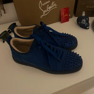 Blå Christian Louboutin sneakers med nitar - Säljer ett par blå sneakers från Christian Louboutin med coola nitar på tån. Skorna har snörning och är gjorda i mocka med en platt sula. Perfekta för dig som vill sticka ut med en unik och edgy stil.