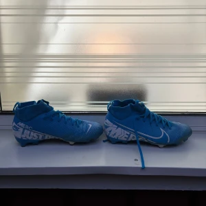 Nike Mercurial blå fotbollsskor - Säljer ett par Nike Mercurial fotbollsskor i klarblå färg med vita detaljer och texten 'JUST DO IT' på sidan. Skorna har snörning, platt sula med dobbar och en strumpliknande högre krage för extra stöd. Materialet är syntet och designen är sportig och modern.