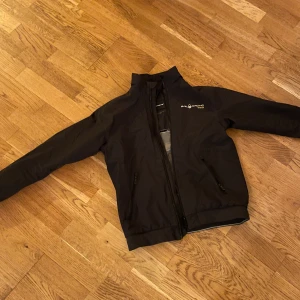 Svart Sail Racing Gore-Tex vind/regnjacka - Svart vind/regn jacka från Sail Racing med Gore-Tex-material, perfekt för blåsiga dagar. Jackan har sidofickor med dragkedja och snyggt broderad logga på bröstet. Stilren design med elastisk nederkant och ärmslut. Storlek 160, Den är 8/10 skick.