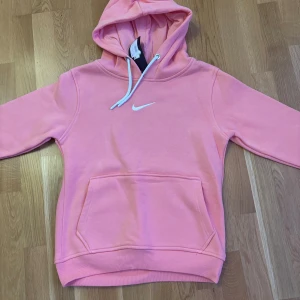 Rosa hoodie från Nike med huva - Säljer en snygg rosa hoodie från Nike i storlek M. Tröjan har en klassisk huva med vita snören, en stor magficka och Nike-logga broderad på bröstet. Mjuk och skön bomullsmix, perfekt för chill eller träning. Långärmad och med ribbade muddar.