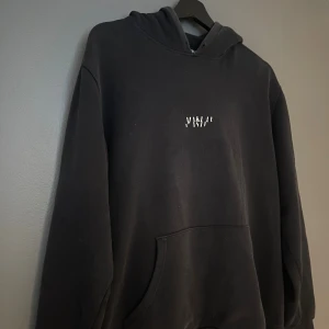 Svart hoodie  - Svart hoodie broderad vit logga på bröstet. Tröjan har huva, stor magficka och långa ärmar. Materialet är mjukt och känns som bomull, perfekt för chill dagar. Passformen är loose och hoodien har en enkel, clean look.