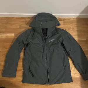 Mörkgrå dunjacka från Arc'teryx - Säljer en mörkgrå dunjacka från Arc'teryx med huva och snyggt broderad logga på bröstet. Jackan har dragkedja framtill, snörning vid fickorna och quiltad insida. Perfekt för kalla dagar och outdoor-äventyr.