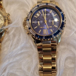 Guldig Michael Kors klocka blå urtavl - Snygg Michael Kors klocka i guldigt metallarmband med blå urtavla och blå detaljer runt boetten. Klockan har romerska siffror, tre mindre urtavlor och datumvisning. Perfekt accessoar för dig som gillar lyxig och stilren design.