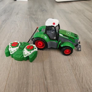 Bull RC Traktor med fjärrkontroll - Fjärrstyrd leksakstraktor från Bull med kabelansluten fjärrkontroll. Grön och svart design med röda hjul, robust konstruktion och tydliga knappar. Produkten är i gott skick utan synliga skador eller slitage. Perfekt för barn som gillar fordon och teknik.