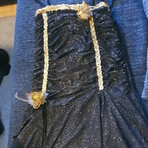 Svart glittrig långklänning med guld - Svart glittrig långklänning med guldiga paljettband och två dekorativa guldrosor. Klänningen har en figurnära passform och är perfekt för speciella tillfällen. Materialet är glittrigt och skimrar snyggt i ljuset.