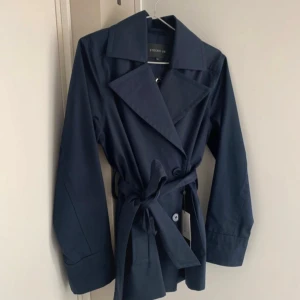 Mörkblå trenchcoat från STOCKH LM XS. Nypris 1700:- - Snygg mörkblå trenchcoat från STOCKH LM i klassisk modell med bred krage, knytskärp i midjan och knappar framtill. Jackan har långa ärmar och stilren design, perfekt för dig som gillar minimalistiskt mode.