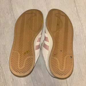Adidas Campus sneakers med rosa ränder - Säljer ett par Adidas Campus sneakers i ljusbeige mocka med klassiska rosa ränder på sidorna och gummisula. Skorna har snörning och rund tå, samt Adidas-logga i guld och rosa på plösen. Perfekta för dig som gillar retrostil och snygga detaljer. Använda 2 gånger. Är större än storlek 36, strax under 37. 