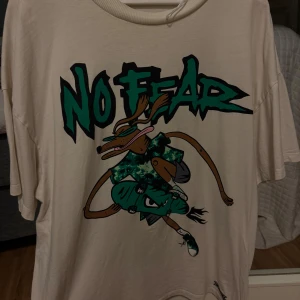 No Fear t-shirt med tryck - Cool vit t-shirt från No Fear med stort grafiskt tryck framtill av en tecknad karaktär och texten 'No Fear' i grönt och svart. T-shirten har korta ärmar och rund hals. 