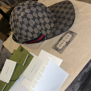 Gucci keps med GG-mönster och röd-blå rand - INGA BYTEN..Säljer en snygg keps från Gucci med klassiskt GG-monogram i mörkblått på beige botten. Kepsen har en röd och marinblå rand på sidan och justerbar rem baktill. Materialet är bomullscanvas med detaljer i läder och tyginsida. Perfekt för dig som vill sticka ut med lyxig streetstyle.