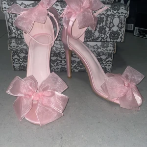 Rosa klackskor med stora rosetter - Snygga ljusrosa pumps med hög klack och öppen tå. Skorna har stora, genomskinliga rosetter både vid ankeln och framtill. Tillverkade i satinliknande material som ger en glansig finish. Perfekta för dig som vill sticka ut med en söt och trendig look.