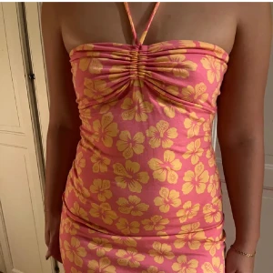 Rosa blommig kortklänning med halterneck - Superfin rosa kortklänning med gula blommor och halterneck. Klänningen har rynkdetalj framtill och är figurnära med ärmlös design. Perfekt för sommarens varma dagar och har en riktigt härlig vibe.