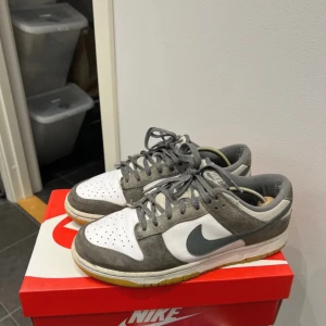 Nike Dunk Low grå/vit EU 42.5 - Nike Dunk Low i grått och vitt med klassisk svart swoosh. Ovandel i mocka och textil, snörning och gummisula i gulbrun ton. Perfekt för dig som gillar streetwear och vill ha en ikonisk sneaker i garderoben. Finns några skavanker på skorna men det är inte som förstör helhetsintrycket.