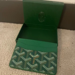 Grön G plånbok med mönster - Snygg grön plånbok från G med det klassiska grafiska mönstret i vitt och brunt. Plånboken har flera kortfack och kommer i original ask. Perfekt för dig som vill ha något unikt och lyxigt i fickan. Plånboken e aldrig andvänd. Nytt skick.