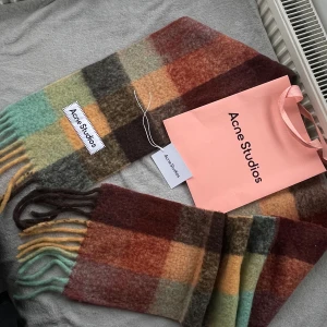 Rutig halsduk Acne Studios ull - Snygg och fluffig halsduk från Acne Studios i rutigt mönster med färger som mintgrön, vinröd, brun, orange och gul. Gjord i mjuk ull, med fransar i ändarna och stor Acne Studios-label. Perfekt för att ge din outfit en cool touch.