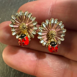 Örhängen med blomma och nyckelpiga i 18k guldpläterad  - Superfina örhängen i 18k guldpläterad med detaljrika blommor i vitt och ljusrosa, och en röd nyckelpiga med svarta prickar på varje örhänge. Perfekt för dig som gillar söta och unika accessoarer med naturtema.