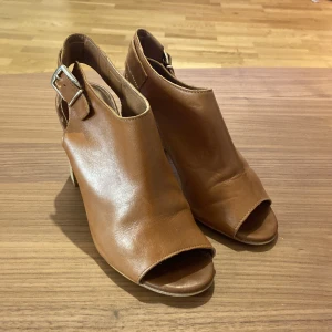 Bruna klackskor Carvela Kurt Geiger - Snygga bruna klackskor från Carvela Kurt Geiger med öppen tå och bred klack i trälook. Skorna har ett stilrent spänne vid hälen och är tillverkade i mjukt skinn. Otroligt sköna klackar.