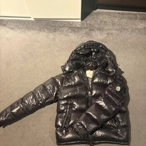 Svart Moncler pufferjacka med huva - Säljer en svart pufferjacka från Moncler med glansig finish och huva. Perfekt för kalla dagar och har snygga detaljer som quiltad design.   Skick 8/10. Fungerande NFC. Storlek 2 som motsvarar S/M. Hör av er vid frågor. 