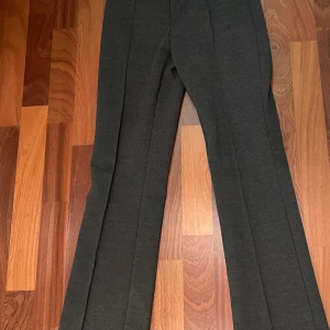 Mörkgrå kostymbyxor med bootcut - Snygga mörkgrå kostymbyxor med bootcut-snitt och pressveck längs benen. Byxorna har en klassisk och stilren look, perfekta för dig som gillar en lite vidare passform nedtill. Materialet känns mjukt och har en lätt stretch för extra komfort.