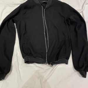 Svart mesh bomberjacka med dragkedja - Svart mesh bomberjacka med klassisk ribbad krage och muddar. Jackan har en silkeslen yta, två sidofickor och en snygg silverfärgad dragkedja framtill. Perfekt för dig som gillar stilren och enkel design med en modern touch.