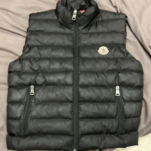 Svart dunväst från Moncler - Svart dunväst från Moncler med quiltad design och hög krage. Västen har två dragkedjeförsedda fickor framtill och ett broderat Moncler-märke på bröstet. Materialet är glansigt och känns lyxigt, perfekt för att lyfta din outfit. Stängs med dragkedja framtill.
