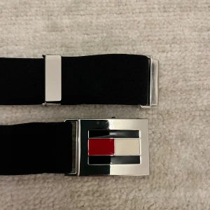 Tommy Hilfiger svart elastiskt bälte - Snyggt svart elastiskt bälte från Tommy Hilfiger med en silverfärgad spänne prydd med det klassiska Hilfiger-flaggan i rött, vitt och mörkblått. Bältet har en stilren och modern look som passar perfekt till jeans och streetwear.