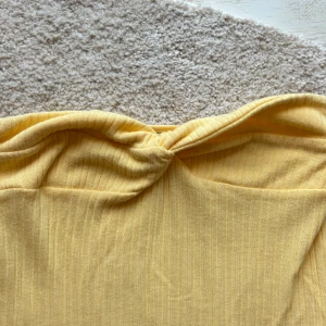 Gul ribbad bandeau tubklänning - Säljer en ljusgul bandeau tubklänning med ribbad struktur och twistad detalj framtill. Toppen är axelbandslös och har en mjuk, stretchig passform som sitter snyggt mot kroppen. Perfekt för varma dagar och enkel att styla med dina favoritjeans eller kjol.