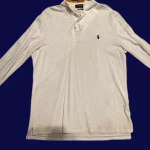 Vit långärmad pikétröja Polo Ralph Lauren - Vit långärmad pikétröja från Polo Ralph Lauren med klassisk krage och knappslå. Tröjan har den ikoniska blå logon broderad på bröstet och är tillverkad i mjuk bomull. Perfekt för dig som gillar stilren och enkel design.