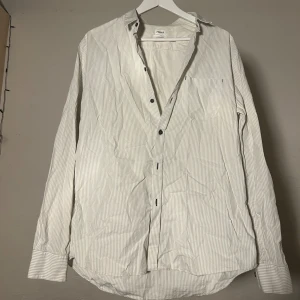 Vit randig skjorta från Filippa K - Vit skjorta från Filippa K med tunna ljusgrå ränder, klassisk krage och svarta knappar. Långärmad modell i mjuk bomull med bröstficka och avslappnad passform. Perfekt för en clean och stilren look. Storlek xl men passar l också.