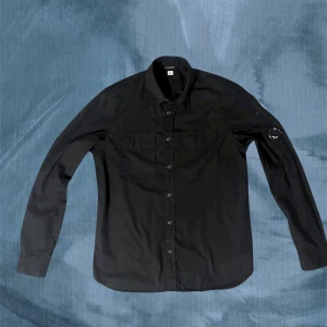 Svart Overshirt C.P. Company - Snygg svart Overshirt från C.P. Company med klassisk krage, två bröstfickor med lock och knappar hela vägen framtill. Ikonisk Stone Island-logga på vänster ärm. Skjortan har en rak passform och är tillverkad i bomull. Linsen och skjortan är som ny, bara använd under speciella tillfällen. Stryks vid beställning 🤩. Nypris på dessa varierar mellan 2500 till 4000 kr.