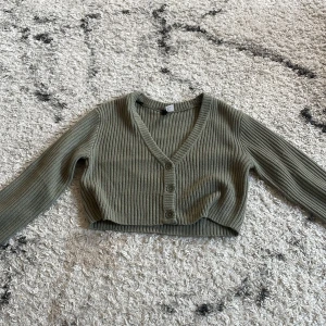 Olivgrön croppad kofta från H&M Divided - Snygg olivgrön croppad kofta från H&M Divided med ribbad struktur och v-ringning. Koftan har tre knappar framtill och långa ärmar. 