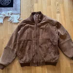 Brun mocka bomberjacka med dragkedja - Snygg brun bomberjacka i mocka med dragkedja framtill och två snedställda fickor. Jackan har ribbade muddar vid ärmslut och midja samt ett mjukt innerfoder. Perfekt för dig som vill ha en stilren och tidlös look. Finns några fläckar, se bilder!