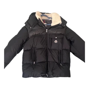 Svart Moncler pufferjacka med huva - Svart pufferjacka från Moncler med huva och mjukt teddyfoder i kragen. Jackan har flera dragkedjor, en bröstficka med Moncler-logga och paneler i skinnimitation upptill. Perfekt för kalla dagar och har en riktigt schysst streetstil.