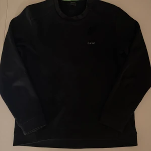 Svart sweatshirt från BOSS - Svart långärmad sweatshirt från BOSS med diskret logga på bröstet och rund hals. Tröjan har en klassisk, clean look och är tillverkad i mjuk bomull med ribbade muddar vid ärmslut och nederkant. Perfekt för chill dagar eller när du vill ha en enkel stil. Använd 1 gång pågrund av liten storlek, nypris 1700 kr och pris kan diskuteras vid snabb affär!!!