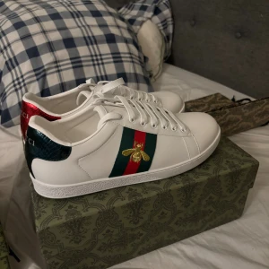 Gucci Ace vita sneakers med bi-detalj - Gucci Ace sneakers i vitt skinn med klassiska gröna och röda ränder på sidan och broderad guldfärgad bi. Baksidan har metallicröd och metallicgrön häl med ormskinnsmönster och Gucci-logga. Platt vit sula och snörning framtill. Riktigt ikoniska och stilrena sneakers.
