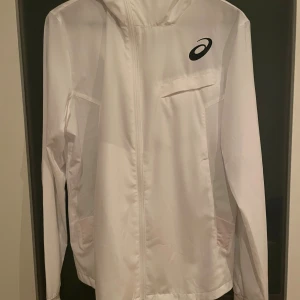 Vit vindjacka från Asics med huva - Snygg vit vindjacka från Asics med huva och hel dragkedja framtill. Jackan har en stilren design med svart Asics-logga på bröstet, praktiska fickor och långärmad passform. Perfekt för dig som gillar sportig och clean stil.