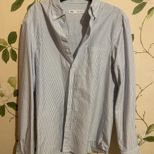 Randig skjorta från Zara, blå/vit - Säljer en klassisk blå- och vitrandig skjorta från Zara i storlek S. Skjortan har lång ärm, bröstficka och knappar framtill. Tillverkad i mjuk bomull och passar perfekt för en clean och stilren look.