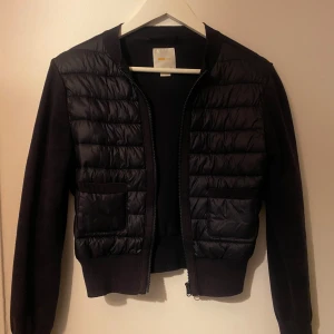 Svart quiltad bomberjacka med dragkedja - Svart bomberjacka från Gina Tricot med quiltad framsida i syntetmaterial och stickade ärmar samt rygg. Jackan har två fickor fram och stängs med dragkedja. Perfekt för dig som gillar en clean och stilren look.