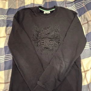 Svart sweatshirt från Kenzo med tiger - Svart sweatshirt från Kenzo i storlek L med broderad tiger och logga på bröstet. Tröjan har rund hals, långa ärmar och är gjord i mjuk bomull. Perfekt för dig som gillar streetwear och vill ha något stilrent med cool detalj framtill.