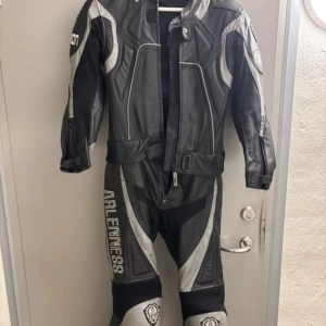 Arlen Ness svart/grå skinnställ strl 38 - Säljer ett racing skinnställ från Arlen Ness i svart och grått med coola detaljer och loggor. Stället har dragkedja framtill, förstärkta partier och snygga vita tryck på ben och axel. Materialet är äkta skinn med polyesterfoder. Perfekt för dig som gillar fart och motorcykel.