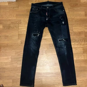 DSQUARED2 mörkblå jeans med slitningar - Säljer ett par mörkblå jeans från DSQUARED2 med coola slitningar, patchdetaljer och färgstänk framtill. Klassisk femficksmodell med gul DSQUARED2-logga baktill i midjan. Jeansen har en rak passform och är tillverkade i jeansmaterial.
