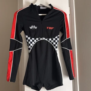 Race-inspirerad dräkt  - Sportig kort dräkt från THATSSOFETCH med svarta och röda paneler, vita detaljer och schackrutigt mönster över midjan. Långärmad med dragkedja framtill och tryckta racingflaggor samt TSF-logga. Tight passform och coolt statement-plagg för dig som gillar motorsport vibes. (Kostar som ny med frakt, tull, moms ca 1400kr)🩷därför lite dyrare 