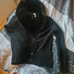Svart ribbad kofta med fuskpäls-krage - Svart ribbstickad kofta från Bodyflirt Boutique med bred, fluffig fuskpäls-krage. Långärmad och knäpps med svarta knappar framtill. Snygg, lite lyxig känsla och passar perfekt när du vill sticka ut. Extra knappar medföljer till den saknade som sista bilden visar!