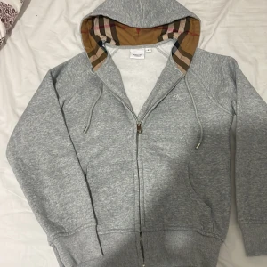 Grå hoodie från Burberry med dragkedja - Grå hoodie från Burberry med klassiskt rutigt foder i huvan och broderad logga på bröstet. Tröjan har snörning i huvan, två fickor fram och dragkedja. Tillverkad i mjuk bomull med ribbade muddar och detaljer i elastan för extra komfort.