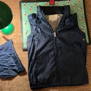 Mörkblå väst från Gucci med GG-mönster - Gucci påsen som man kan lägga in västen. Snygg mörkblå väst från Gucci med ikoniskt GG-mönster över hela tyget. Västen har dragkedja framtill, ståkrage och grått innerfoder med en praktisk innerficka. Perfekt för dig som vill ha en exklusiv och stilren look. Finns en luva att fälla ut! Den är för tolv och 13 år, en jävligt fet väst. Har påse till om man vill ha den är och Gucci!