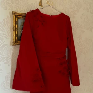 Röd långklänning med utsvängda ärmar - Snygg röd långklänning med utsvängda långa ärmar och dekorativa blommor på axel, ärm och längs sidorna. Klänningen har en elegant, figurnära passform och rund halsringning. Perfekt för dig som vill sticka ut med en unik och färgstark look.