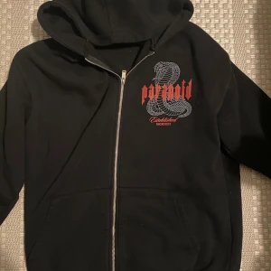 Svart hoodie med tryck från Black Squad - Svart hoodie från Black Squad i relaxed fit med dragkedja och huva. På bröstet finns ett stort tryck med en orm och texten 'paranoid' i rött. Tröjan har fickor framtill och är tillverkad i mjuk bomull. Perfekt för dig som gillar streetwear och coola detaljer.