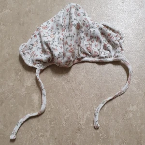 Mössa - Supersöt mössa från Newbie i mjuk bomull med blommigt mönster i rosa och grönt på vit botten. Mössan har praktiska knytband under hakan och en lätt rynkad form som ger en gullig look. Perfekt för dig som gillar romantiska detaljer och mjuka material.