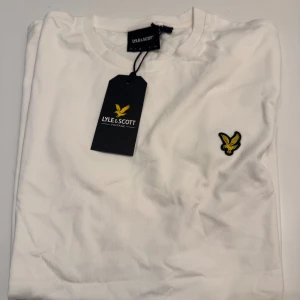 Vit t-shirt från Lyle & Scott - Vit t-shirt från Lyle & Scott med klassisk rund hals och korta ärmar. På bröstet sitter den ikoniska gula örnloggan som broderad detalj. T-shirten är gjord i mjuk bomull och har en clean, stilren look som funkar till allt.                               Pris kan diskuteras 😊
