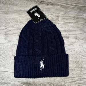 Marinblå kabelstickad mössa Polo - Snygg marinblå mössa från Polo Ralph Lauren med klassisk kabelstickad design och uppvikt kant. Framtill syns den ikoniska vita Polo-loggan broderad. Perfekt för dig som vill ha en stilren och varm accessoar i vinter. Ete kvalité 