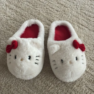 Hello Kitty fluffiga tofflor - Supergulliga vita tofflor med Hello Kitty-design, mjuk pälsimitation och röda rosetter framtill. Insidan är röd och fluffig, perfekt för att hålla fötterna varma hemma. Rund tå och platt sula för extra komfort.